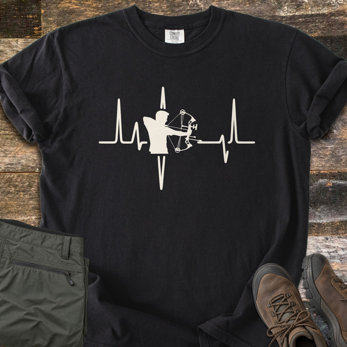 Compound Bow Heartbeat Comfort Colors T-shirt in Black, laid flat. A cream heartbeat line with the silhouette of an archer drawing a compound bow at its center. Sleeves are rolled on a wood plank floor, with olive cargo pants and brown hiking boots beside it.