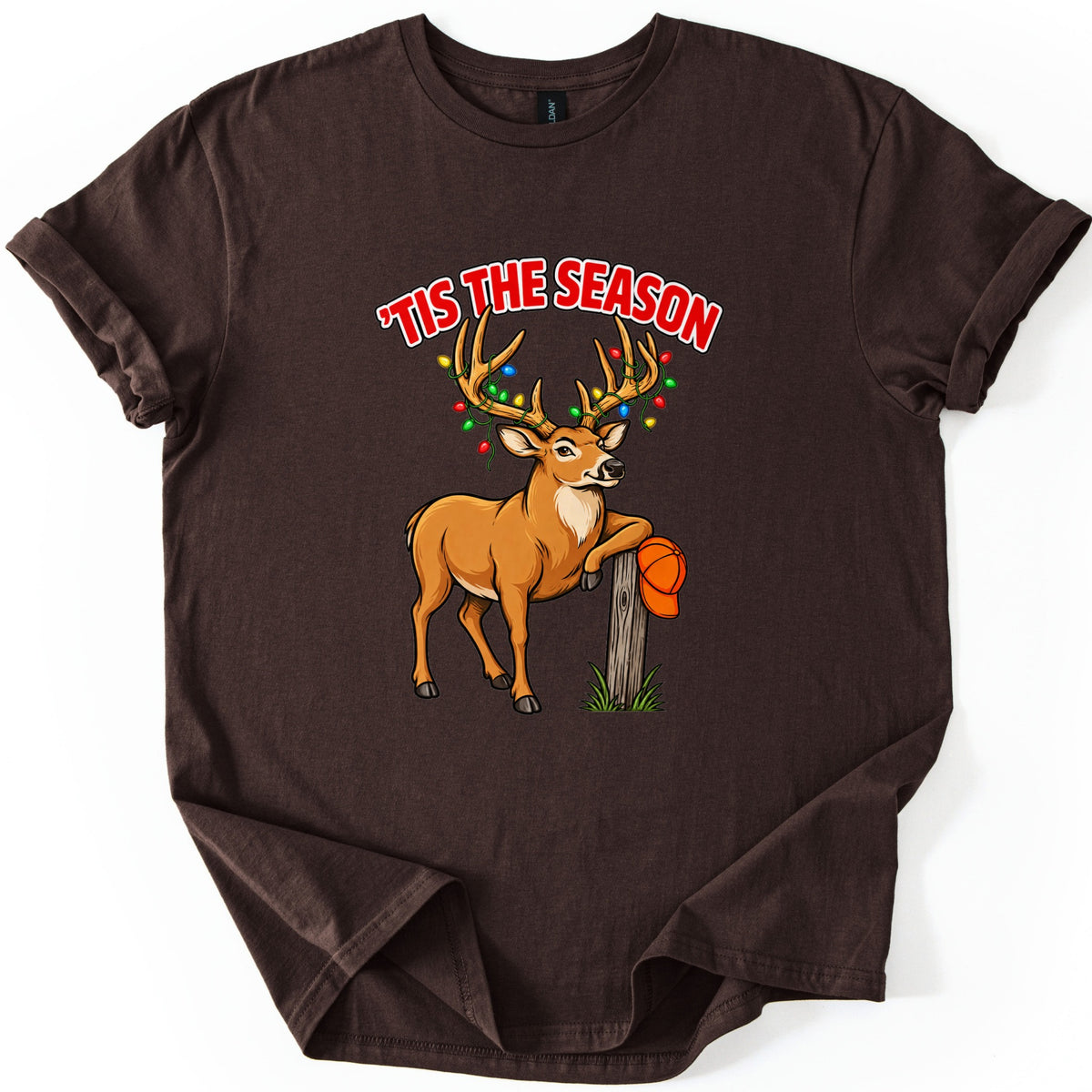 Buck Tis The Season Comfort Colors T-shirt in Dark Chocolate, laid flat. Red arched text reads 'TIS THE SEASON above a cartoon buck with colored Christmas lights strung on its antlers, leaning on a wooden post that holds an orange hunting cap. Sleeves rolled, on a white surface.