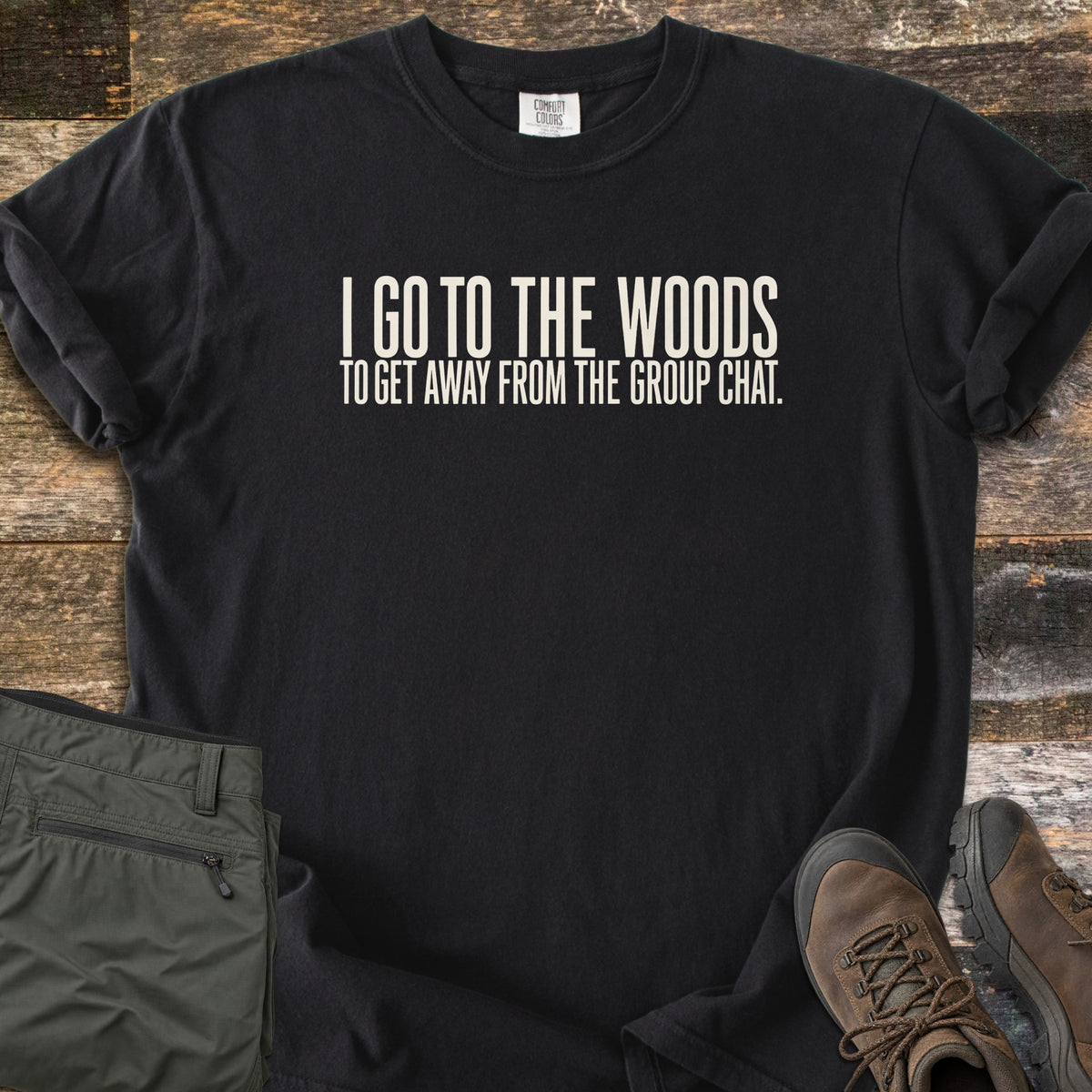 Away From The Group Chat Comfort Colors T-shirt in Black, laid flat. Off-white block letters read I GO TO THE WOODS above TO GET AWAY FROM THE GROUP CHAT. The shirt has rolled sleeves and lies on weathered wood boards beside folded olive green pants and brown hiking boots.