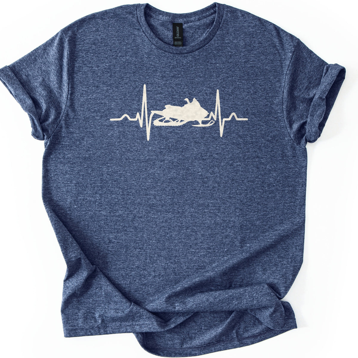 Heartbeat Snowmobile Comfort Colors T-shirt in Heather Navy, laid flat. A cream heartbeat line runs across the chest with a snowmobile silhouette set into the middle of the pulse, with no words. The shirt has rolled sleeves and lies on a white background.