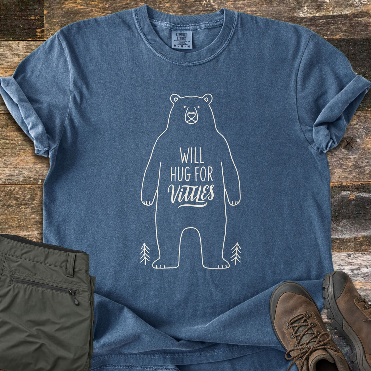 Will Hug For Vittles Comfort Colors T-shirt in Denim, laid flat. A cream outline of a standing bear has the words Will Hug For Vittles on its belly, with two small pine trees by its feet. Folded olive hiking pants and brown hiking boots sit beside it on weathered wood.