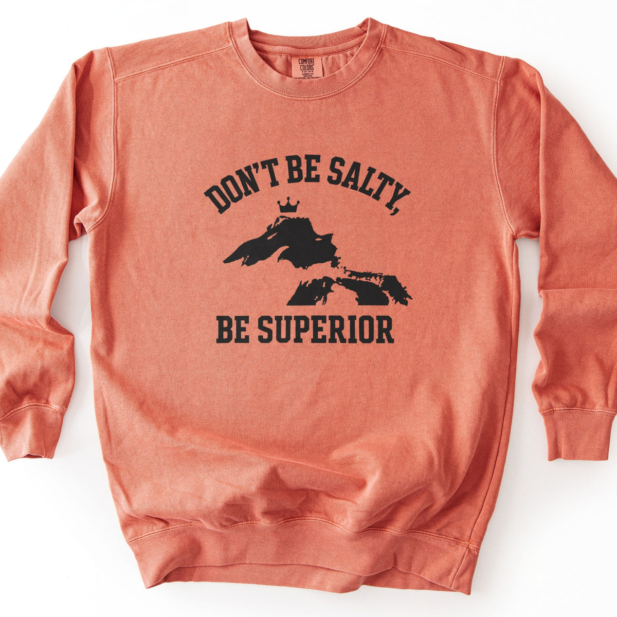 Be Superior Comfort Colors crewneck sweatshirt in Terracotta, laid flat. Black print of the words Don't Be Salty, arched above a silhouette of Lake Superior topped with a small crown, with Be Superior below. Laid on a white background.