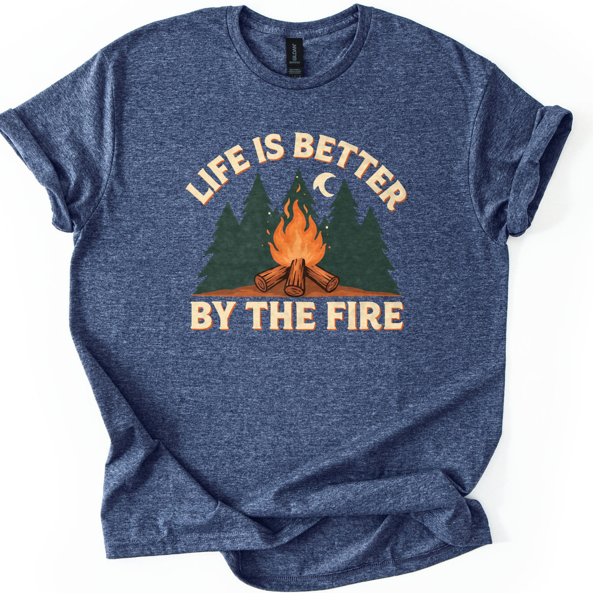 Life Is Better By The Fire Comfort Colors T-shirt in Heather Navy, laid flat. A campfire of crossed logs burns in front of dark green pine trees under a crescent moon, with LIFE IS BETTER arched above and BY THE FIRE below in cream letters. Cuffed sleeves, white background.