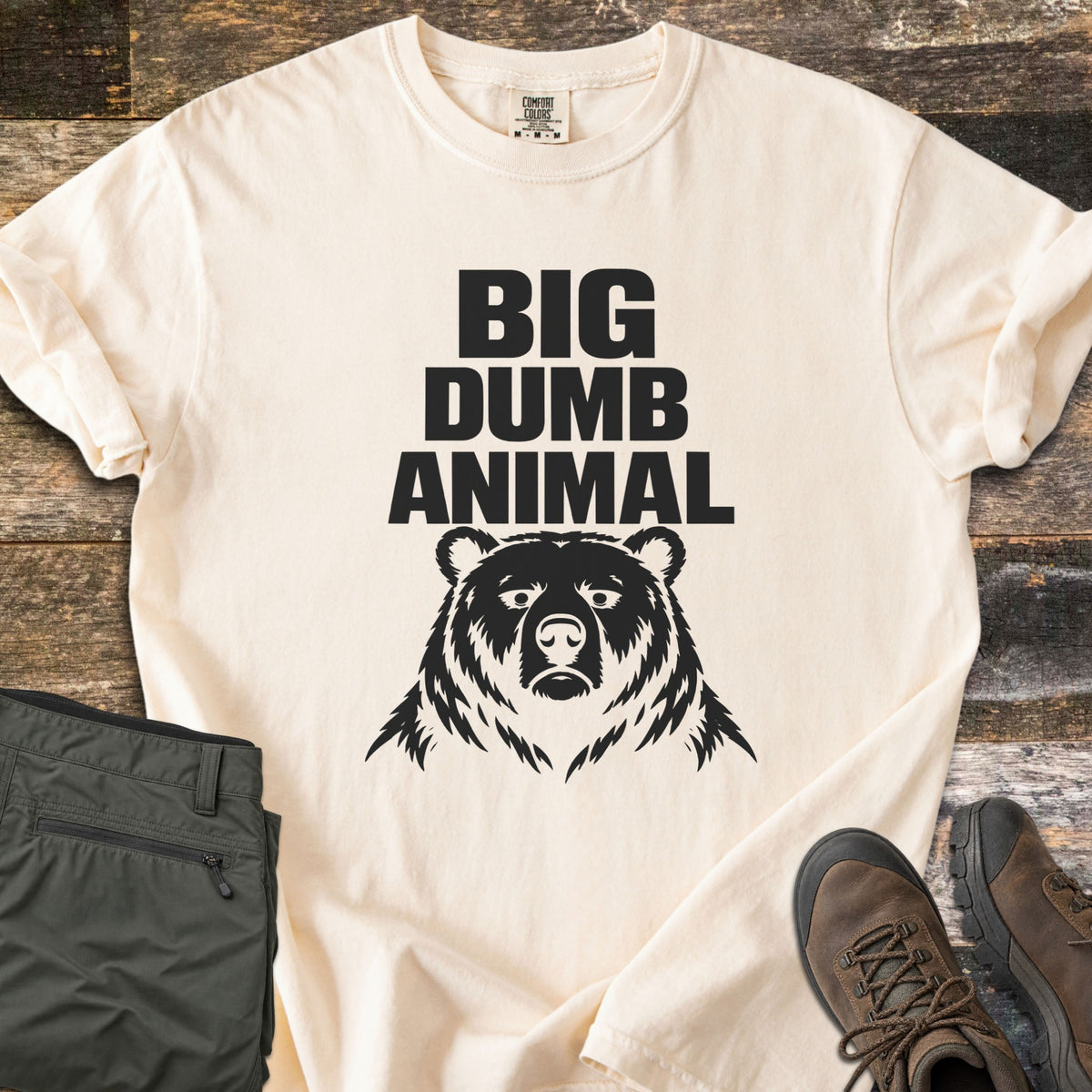 Big Dumb Animal Comfort Colors T-shirt in Ivory, laid flat. Bold black block letters read Big Dumb Animal above a grumpy-faced bear head. The shirt, sleeves rolled, lies on weathered wood planks beside folded olive hiking pants and brown hiking boots.