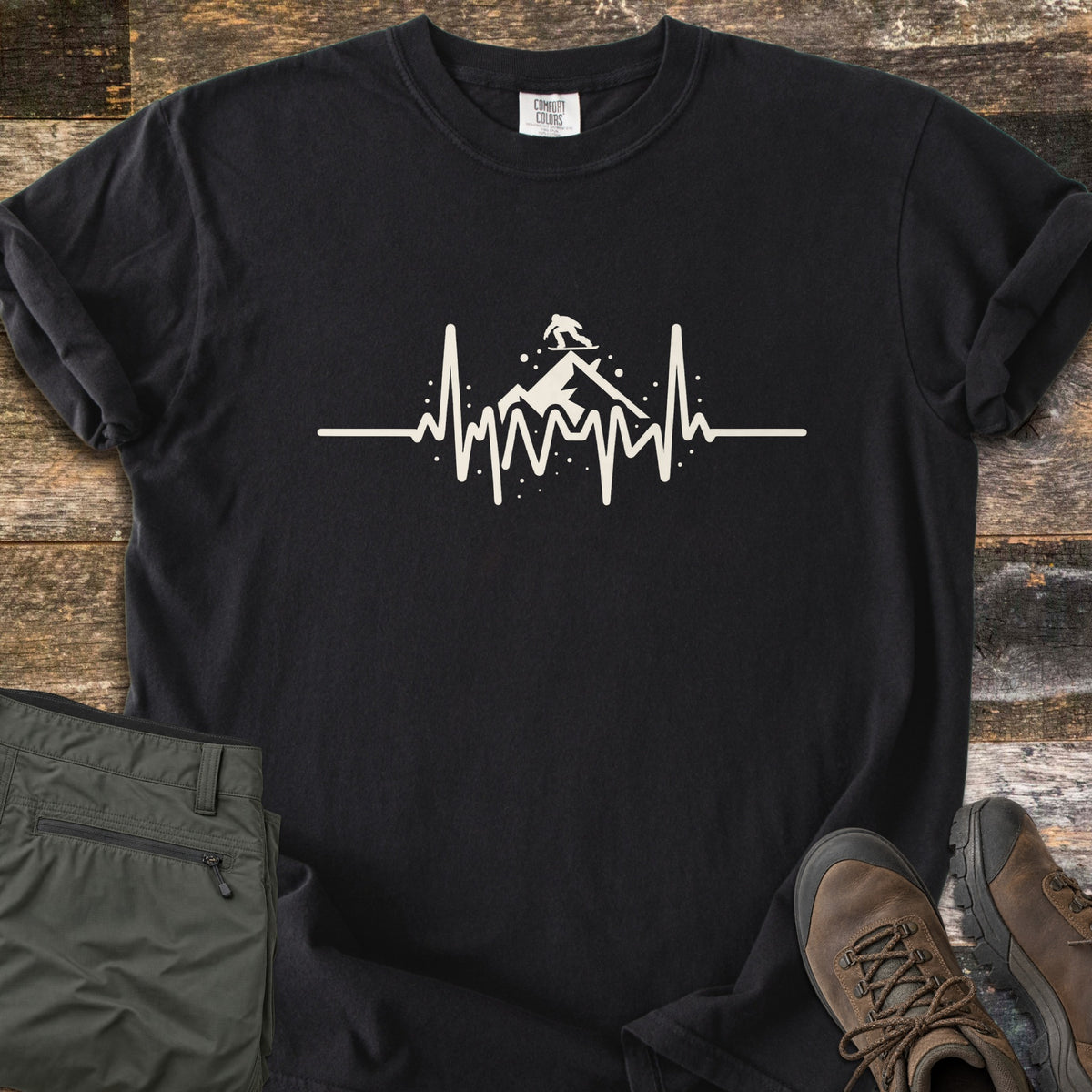 Snow Boarder Heartbeat Comfort Colors T-shirt in Black, laid flat. A cream heartbeat line rises into a mountain peak with a snowboarder riding on top, surrounded by snow dots. Folded olive hiking pants and brown leather boots sit beside it on weathered wood planks.