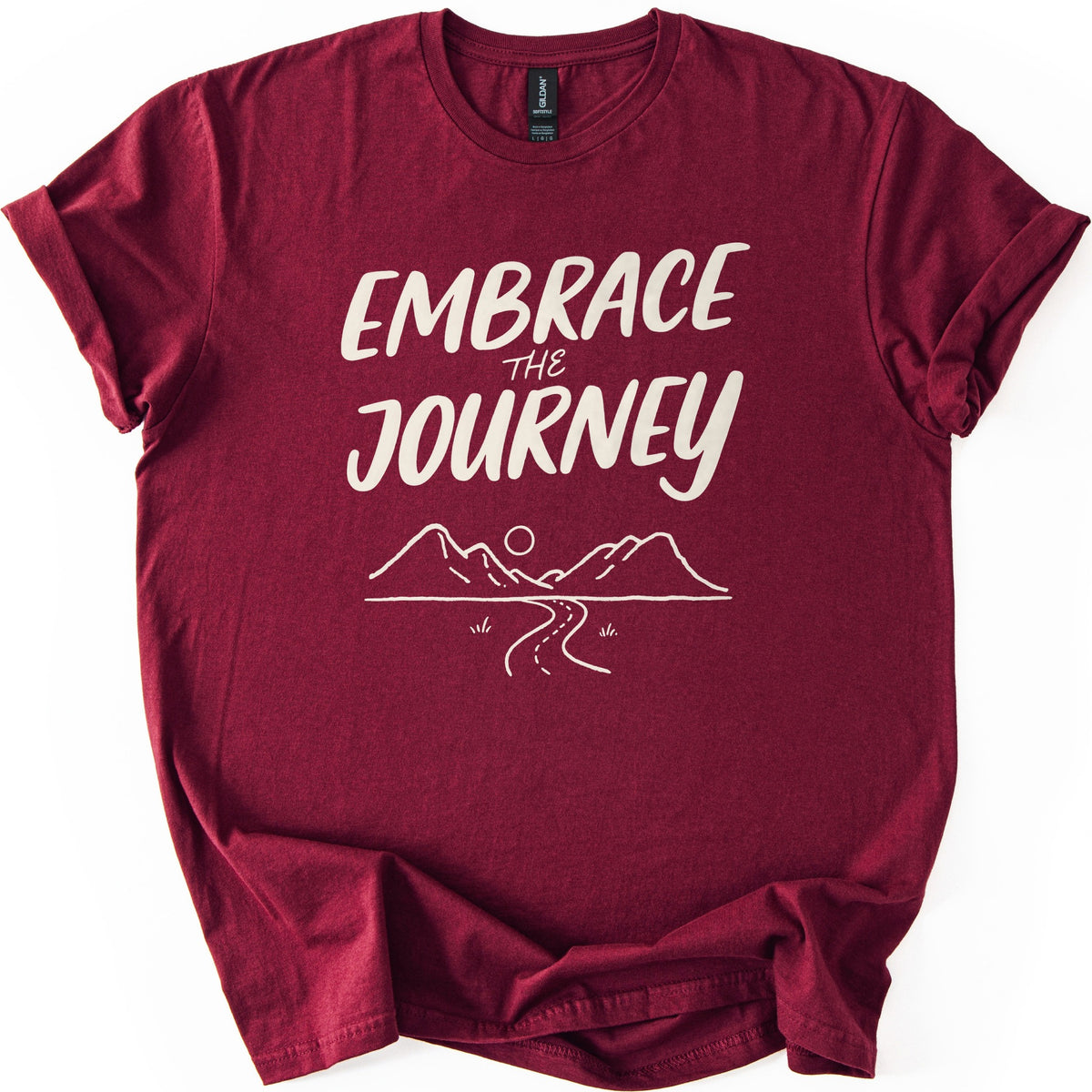 Embrace The Journey Trail Comfort Colors T-shirt in Maroon, laid flat. Cream hand-lettered words Embrace the Journey sit above a line drawing of two mountains, a round sun, and a winding road with tufts of grass. The sleeves are cuffed, on a white background.