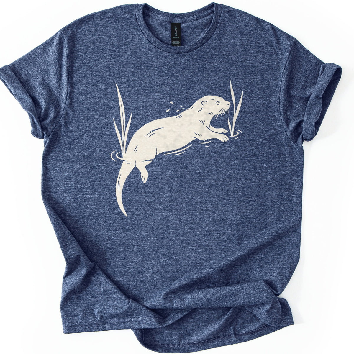 Swimming Otter Comfort Colors T-shirt in Heather Navy, laid flat. A cream linocut-style otter swims through rippling water between tall reeds, with droplets above its head. No words are printed. The sleeves are cuffed and the shirt lies on a white background.
