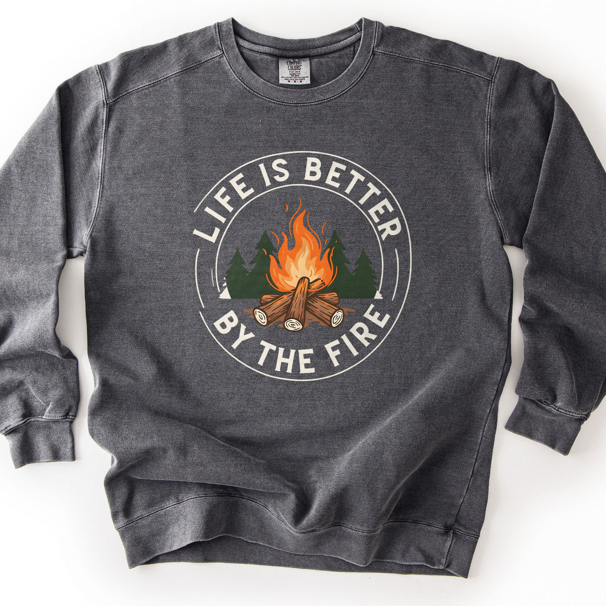 Life Is Better By The Fire Ring Comfort Colors crewneck sweatshirt in Pepper, laid flat. A circular badge shows a campfire of crossed logs with orange flames before dark green pine trees, ringed by the words Life Is Better By The Fire in cream lettering. It lies on a white surface.