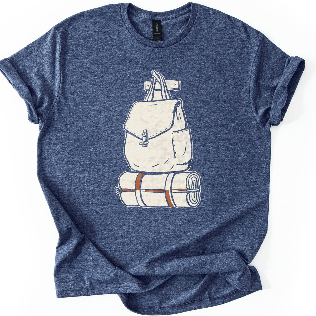 Backpack And Bedroll Comfort Colors T-shirt in Heather Navy, laid flat. A cream illustration of a canvas backpack with a toggle-clasp flap hangs by its handles from a wall peg above a rolled bedroll tied with rust-orange straps. No words. Cuffed sleeves on a white surface.
