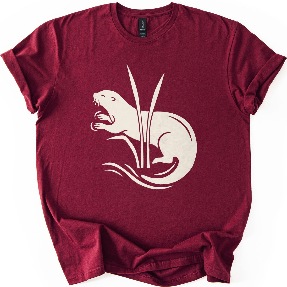 Otter In The Reeds Comfort Colors T-shirt in Maroon, laid flat. A cream silhouette of an otter with open mouth, whiskers and raised paws curls behind two tall reeds above swirling water lines. No words. The shirt lies on a white surface with sleeves rolled.