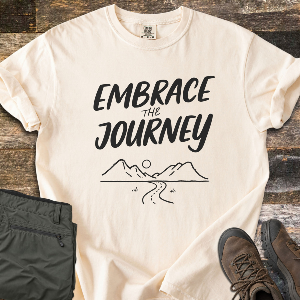 Embrace the Journey Comfort Colors T-shirt in Ivory, laid flat. Black brush lettering reads Embrace the Journey above a line drawing of mountains, a sun, and a winding road. Sleeves cuffed on weathered wood beside folded green pants and brown hiking boots.