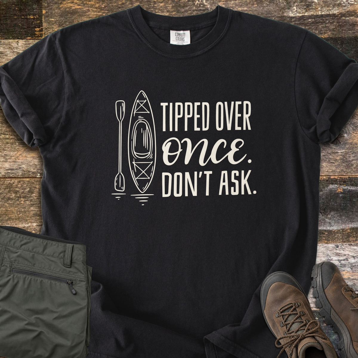 Tipped Over Once. Don't Ask. Comfort Colors T-shirt in Black, laid flat. A white line drawing of a kayak and paddle sits beside the words Tipped Over Once. Don't Ask., with Once in script. Sleeves rolled, on weathered wood with folded olive hiking pants and brown hiking boots.