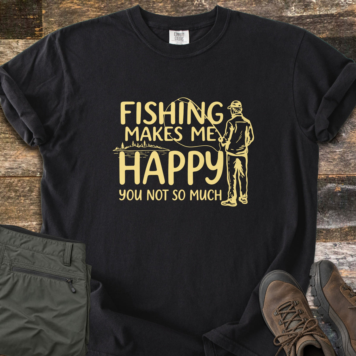 Fishing Makes Me Happy Comfort Colors T-shirt in Black, laid flat. Pale yellow print of a man in a cap casting a fishing rod by a tree-lined lake, with the words Fishing Makes Me Happy You Not So Much. Laid on weathered wood with olive pants and brown hiking boots.