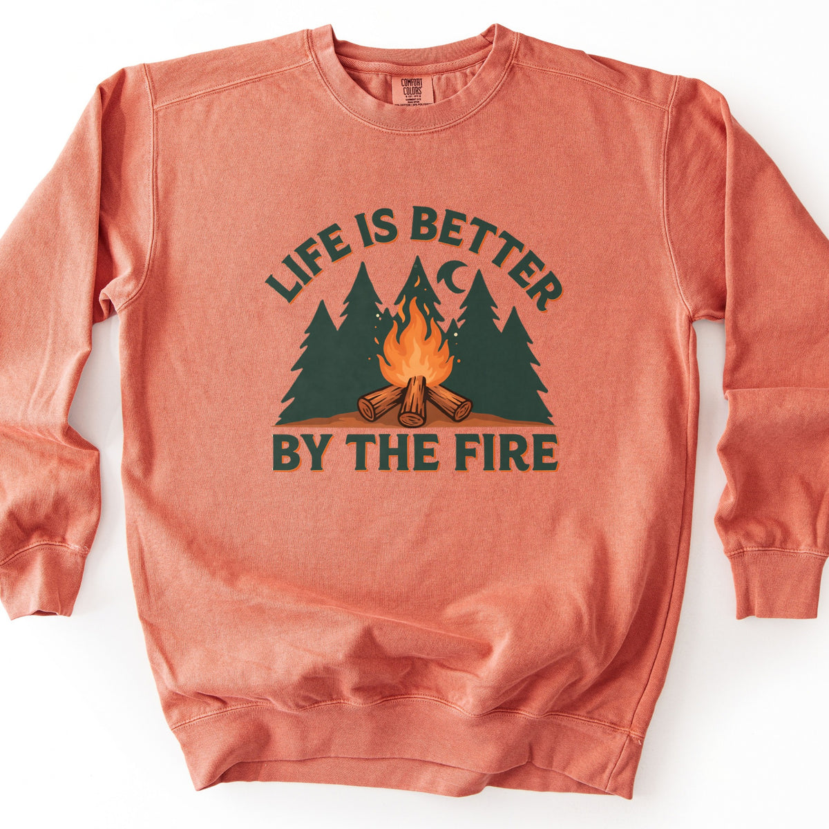 Life Is Better By The Fire Comfort Colors crewneck sweatshirt in Terracotta, laid flat. Dark green letters with an orange shadow arch Life Is Better over dark green pine trees, a crescent moon and a campfire of crossed logs, with By The Fire below. It lies on a white surface.