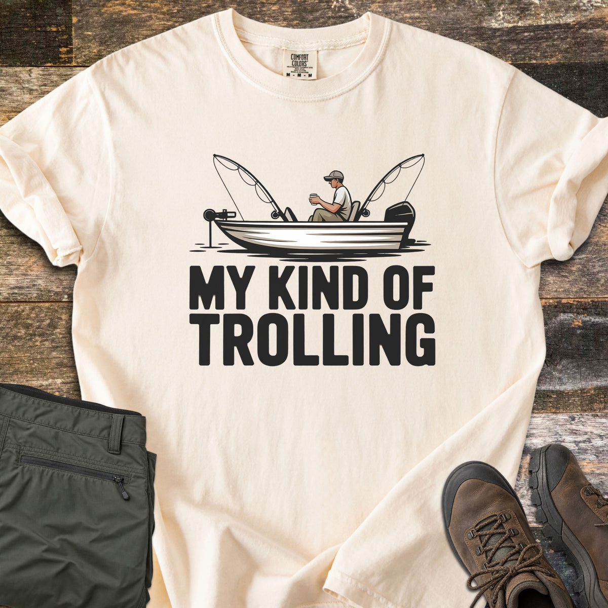 My Kind Of Trolling Comfort Colors T-shirt in Ivory, laid flat. A man in a cap holds a cup in a small fishing boat with two bent rods and motors, above the words My Kind Of Trolling in black. Olive cargo pants and brown hiking boots sit beside it on weathered wood.