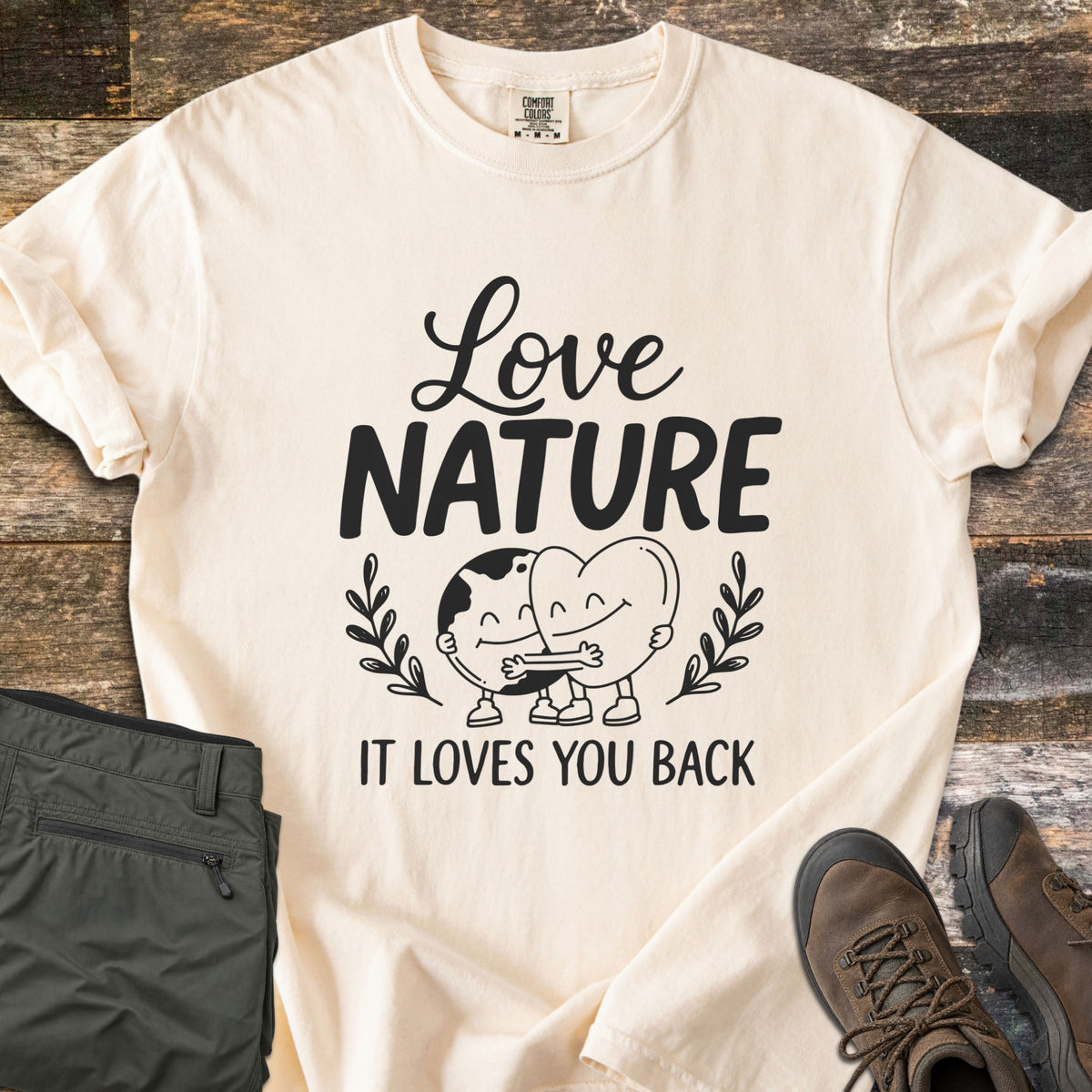 Nature Loves You Back Comfort Colors T-shirt in Ivory, laid flat. Black print reads Love Nature above a smiling cartoon Earth and heart hugging between leafy sprigs, with It Loves You Back below. It lies on weathered wood beside olive hiking pants and brown boots.