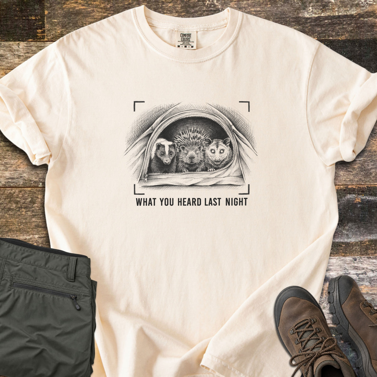 What You Heard Last Night Comfort Colors T-shirt in Ivory, laid flat. A black sketch of a skunk, porcupine and opossum peeking out of an unzipped tent, framed by camera brackets, above the words What You Heard Last Night. Olive hiking pants and brown boots lie on weathered wood.