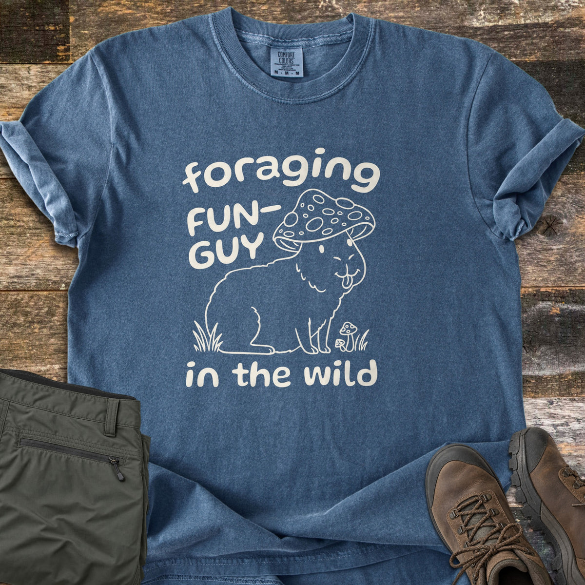 Foraging Fun-Guy in the Wild Comfort Colors T-shirt in Denim, laid flat. A cream line drawing of a capybara wearing a spotted mushroom cap, tongue out, sits in grass by tiny mushrooms between the words foraging FUN-GUY in the wild. Olive hiking pants and brown boots lie on weathered wood.