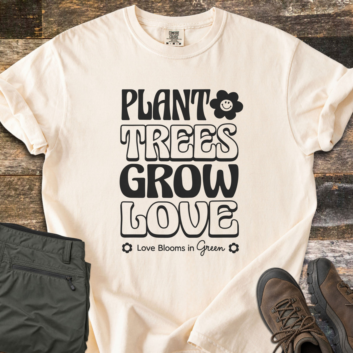 Plant Trees Grow Love Comfort Colors T-shirt in Ivory, laid flat. Black retro bubble letters read Plant Trees Grow Love beside a smiling daisy, above Love Blooms in Green. Sleeves cuffed on weathered wood with folded olive hiking pants and brown hiking boots.