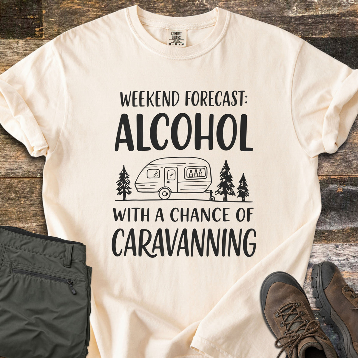 Alcohol With a Chance of Caravanning Comfort Colors T-shirt in Ivory, laid flat. Black print reads Weekend Forecast: Alcohol above a caravan with bottles in its window between pine trees, then With a Chance of Caravanning. Olive trousers and brown hiking boots sit on dark wood planks.