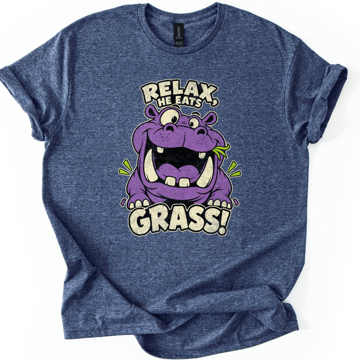 Relax He Eats Grass Comfort Colors T-shirt in Heather Navy, laid flat. A grinning purple cartoon hippo with big tusks and a blade of grass in its mouth sits between the words RELAX, HE EATS and GRASS! in cream letters. The shirt lies on white with rolled sleeves.