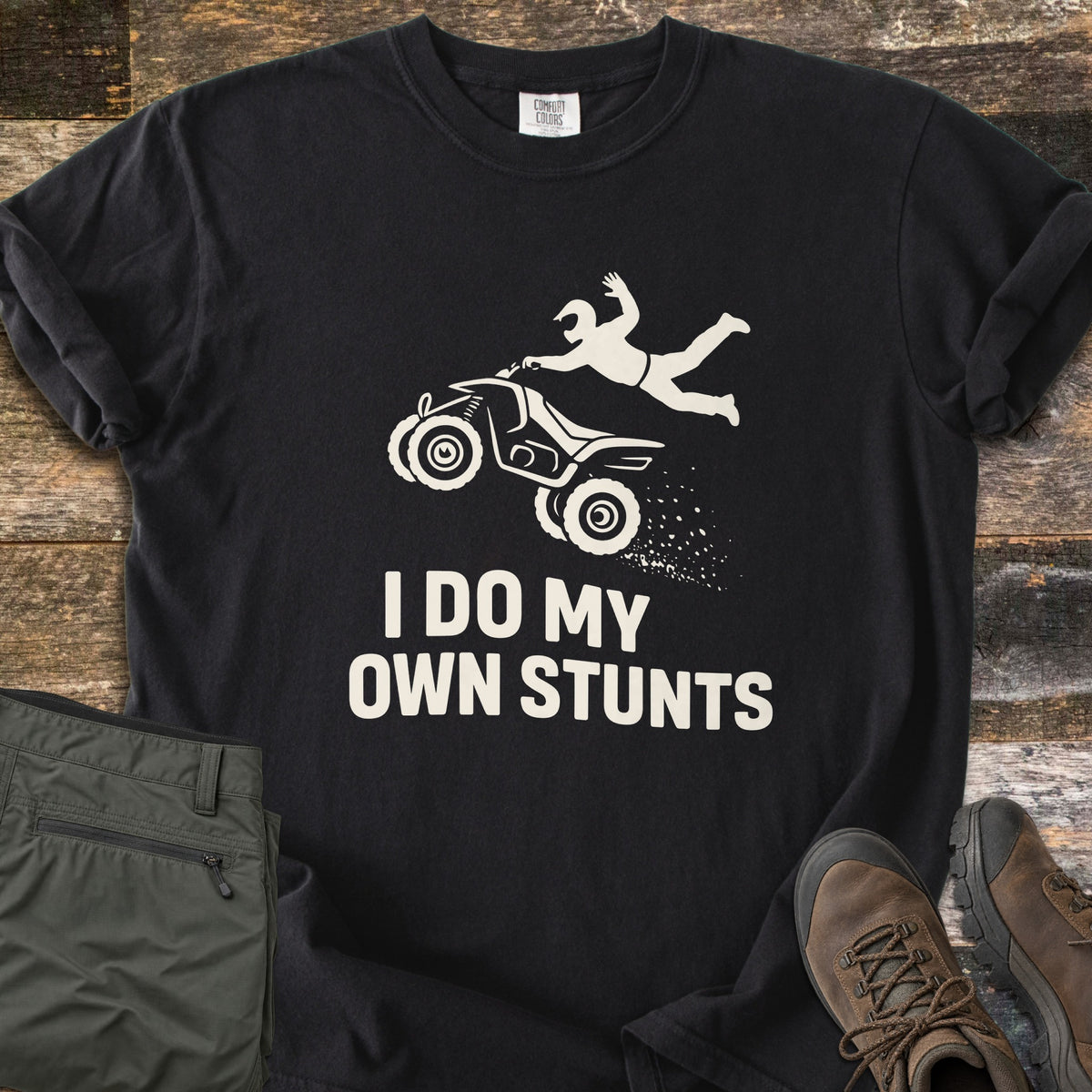 ATV Stunts Comfort Colors T-shirt in Black, laid flat. A white silhouette of a helmeted rider flying off a wheelie-popping ATV kicking up dirt, above the words I Do My Own Stunts. Olive cargo pants and brown hiking boots sit beside it on weathered wood.