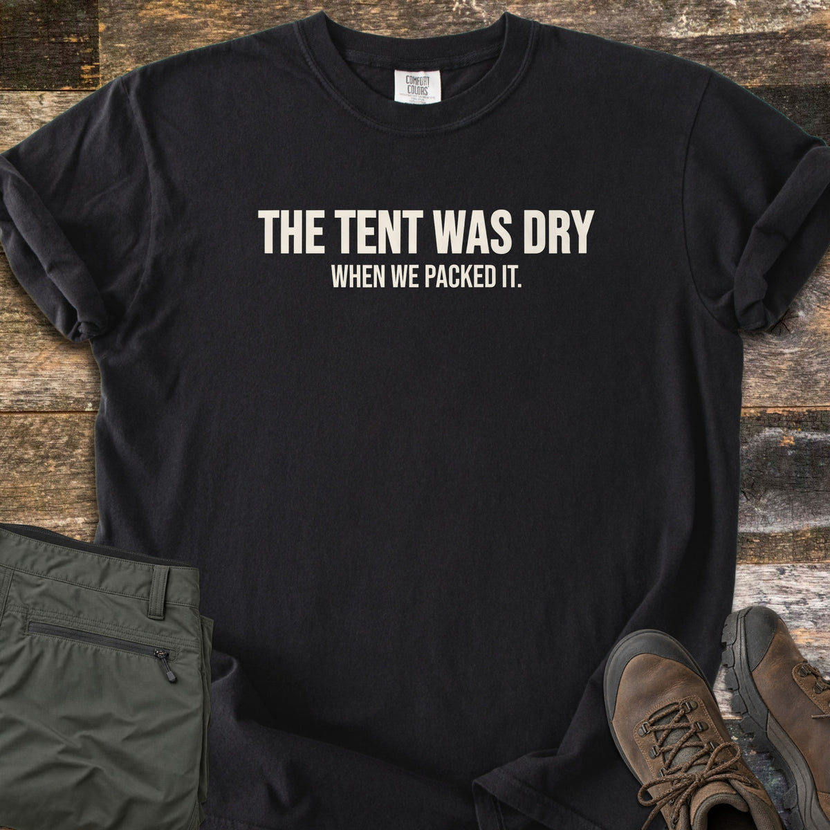 The Tent Was Dry Comfort Colors T-shirt in Black, laid flat. Bold cream capital letters read THE TENT WAS DRY, with smaller text below reading WHEN WE PACKED IT. The shirt lies on weathered wood planks with rolled sleeves, beside folded olive hiking pants and brown hiking boots.