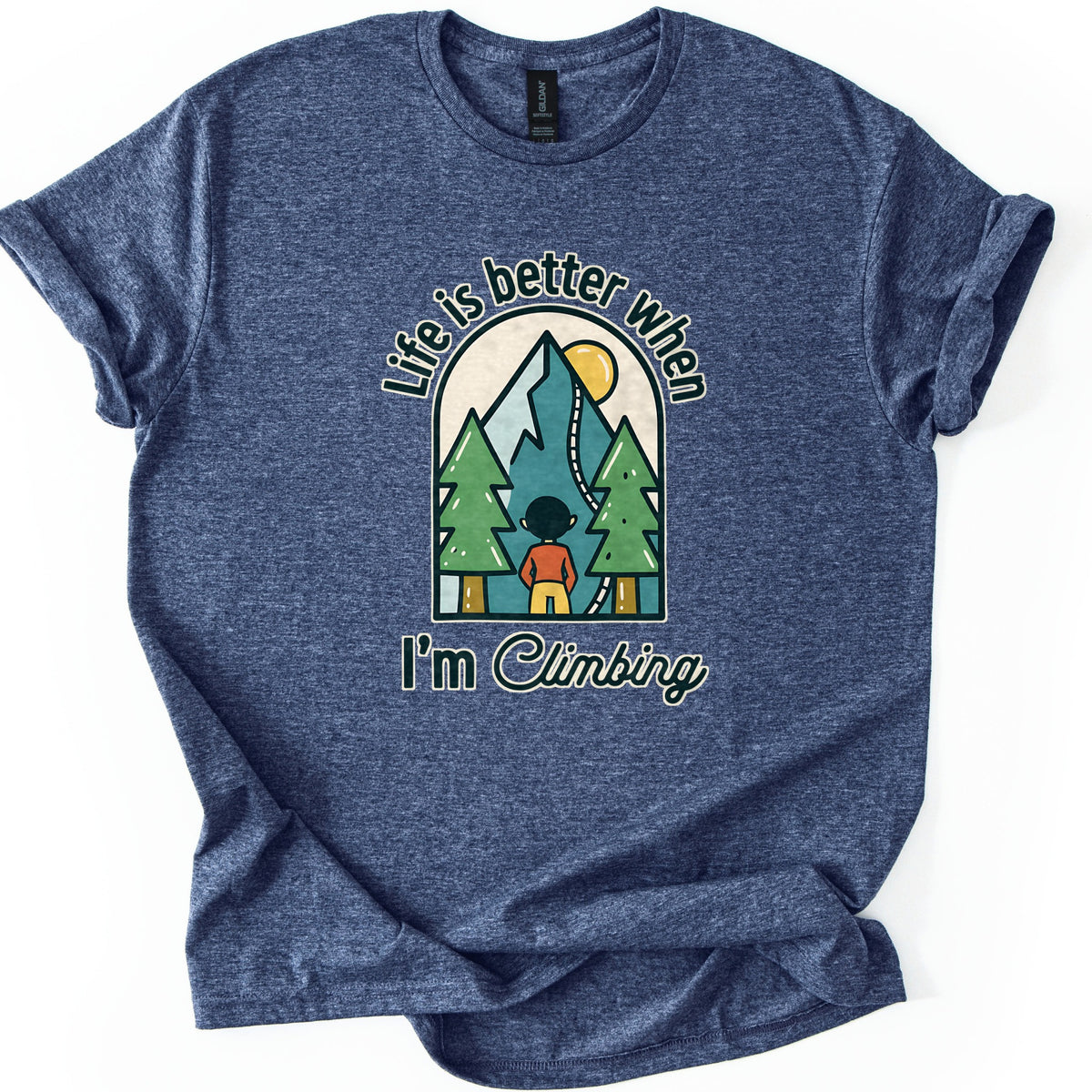 Better When I'm Climbing Comfort Colors T-shirt in Heather Navy, laid flat. The words Life is better when arch over a window scene of a person facing a teal mountain with a winding trail, a yellow sun and two pine trees, with I'm Climbing below. Sleeves cuffed, on a white surface.