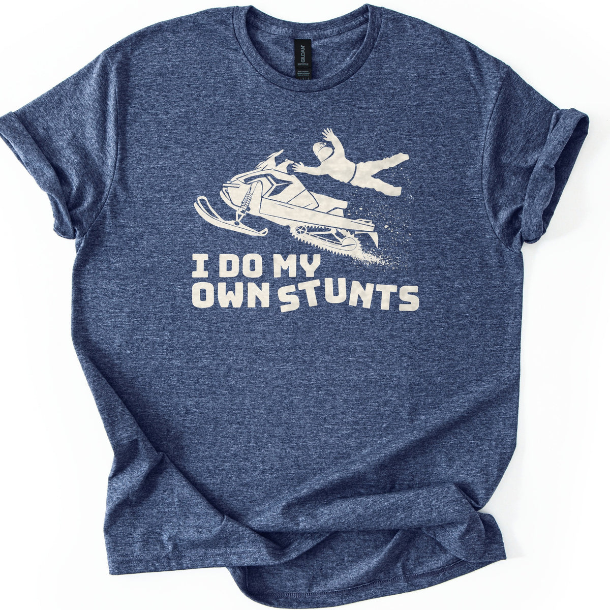 Snow Mobile Stunts Comfort Colors T-shirt in Heather Navy, laid flat. A cream silhouette of a snowmobile flying through the air, its rider thrown off behind it in a spray of snow, above the words I Do My Own Stunts. The shirt lies on a white background with rolled sleeves.