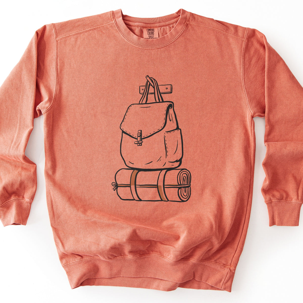 Backpack And Bedroll Comfort Colors crewneck sweatshirt in Terracotta, laid flat. Black line art of a canvas backpack with a toggle clasp hanging by its straps from a wall peg above a rolled bedroll tied with brown straps. No words are printed. Shown on a white background.
