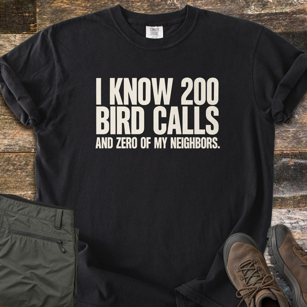 I Know 200 Bird Calls and Zero of My Neighbors Comfort Colors T-shirt in Black, laid flat. Bold off-white capital letters read I KNOW 200 BIRD CALLS AND ZERO OF MY NEIGHBORS. The shirt has rolled sleeves and lies on weathered wood boards beside folded olive hiking pants and brown hiking boots.