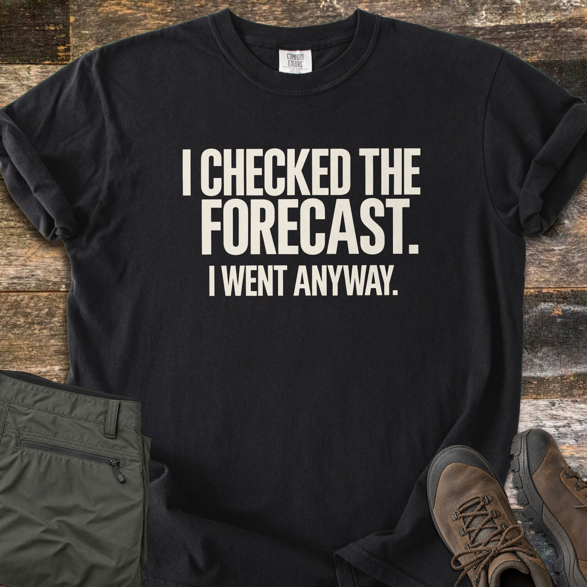 Checked The Forecast Comfort Colors T-shirt in Black, laid flat. Off-white block letters read I CHECKED THE FORECAST. I WENT ANYWAY. The sleeves are cuffed, with folded olive hiking pants and brown hiking boots beside it on weathered wood planks.