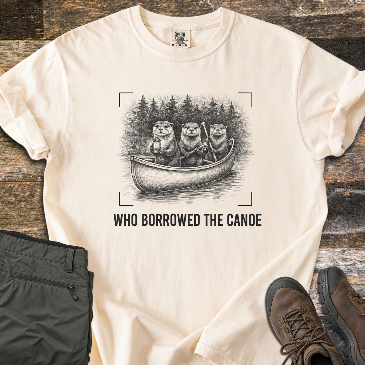 Who Borrowed The Canoe Comfort Colors T-shirt in Ivory, laid flat. A black sketch of three otters in a canoe before pine trees, one holding a lantern, framed by camera corner marks above the words Who Borrowed The Canoe. Olive hiking pants and brown hiking boots sit on dark wood.