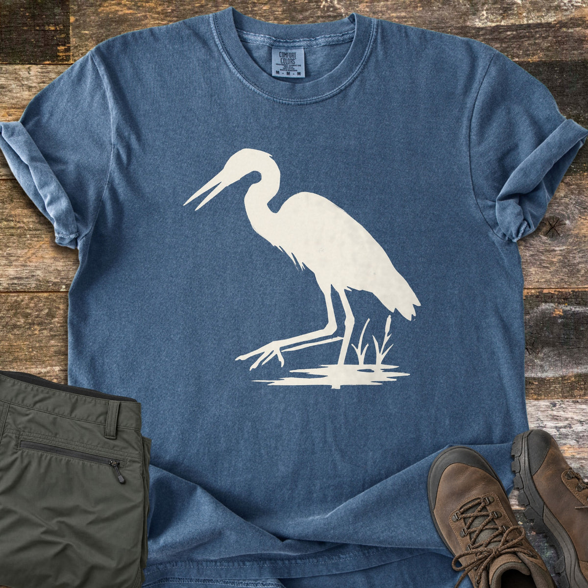 Great Blue Heron Comfort Colors T-shirt in Denim, laid flat. A cream silhouette of a heron wading in shallow water beside reeds, with no words. The shirt lies on weathered wood planks with rolled sleeves, beside olive cargo pants and brown leather hiking boots.