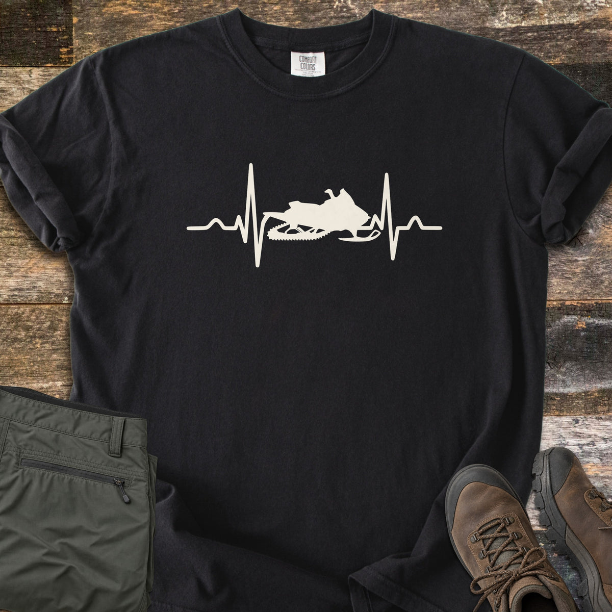 Snowmobile Heartbeat Comfort Colors T-shirt in Black, laid flat. A cream heartbeat line runs across the chest with a snowmobile silhouette in its center, with no words. Sleeves are rolled, on weathered wood planks beside folded olive pants and brown hiking boots.