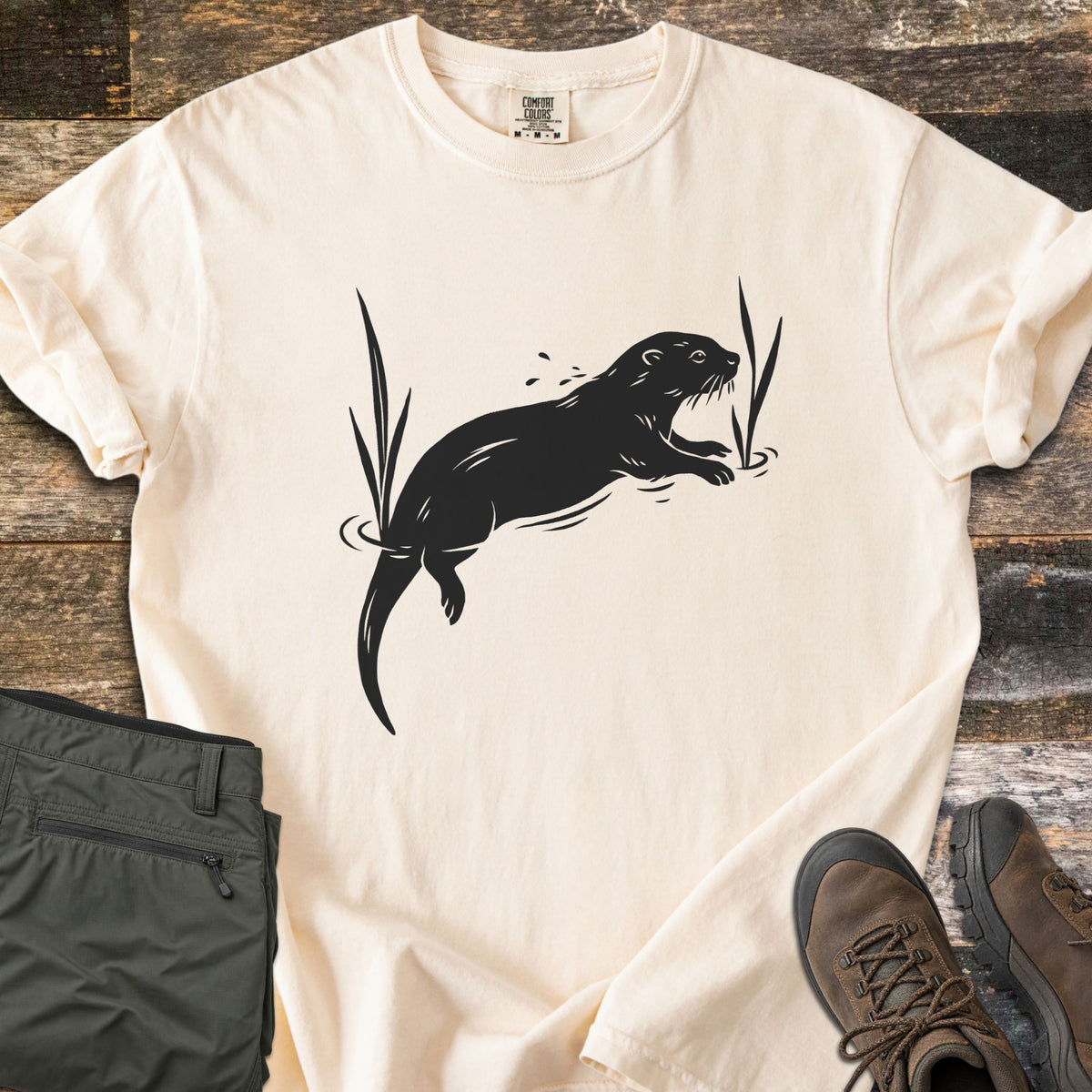 Swimming Otter Comfort Colors T-shirt in Ivory, laid flat. A black print of an otter swimming between tall reeds, with ripples and splashing droplets. Sleeves are rolled; olive hiking pants and brown hiking boots sit on weathered wood planks.