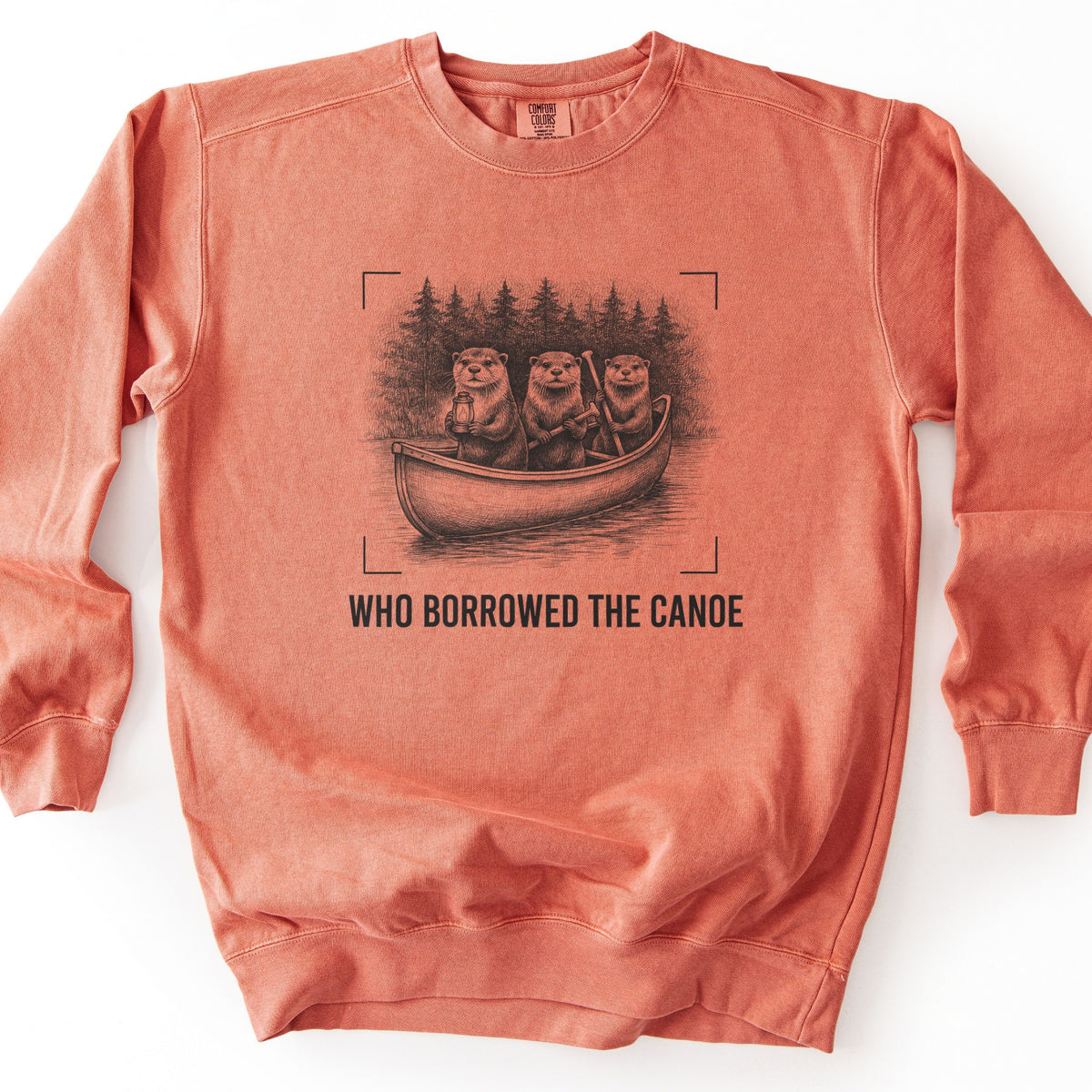 Who Borrowed The Canoe Comfort Colors crewneck sweatshirt in Terracotta, laid flat. A black sketch of three otters in a canoe, one holding a lantern and two gripping paddles, with a pine forest behind and corner brackets around it, above the words WHO BORROWED THE CANOE. On a white surface.