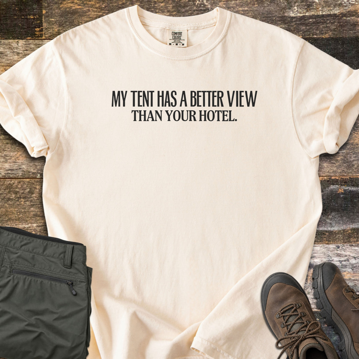 Better View Than Your Hotel Comfort Colors T-shirt in Ivory, laid flat. Black text reads My Tent Has a Better View Than Your Hotel. Sleeves are cuffed, on a weathered wood surface with folded olive hiking pants and brown hiking boots.
