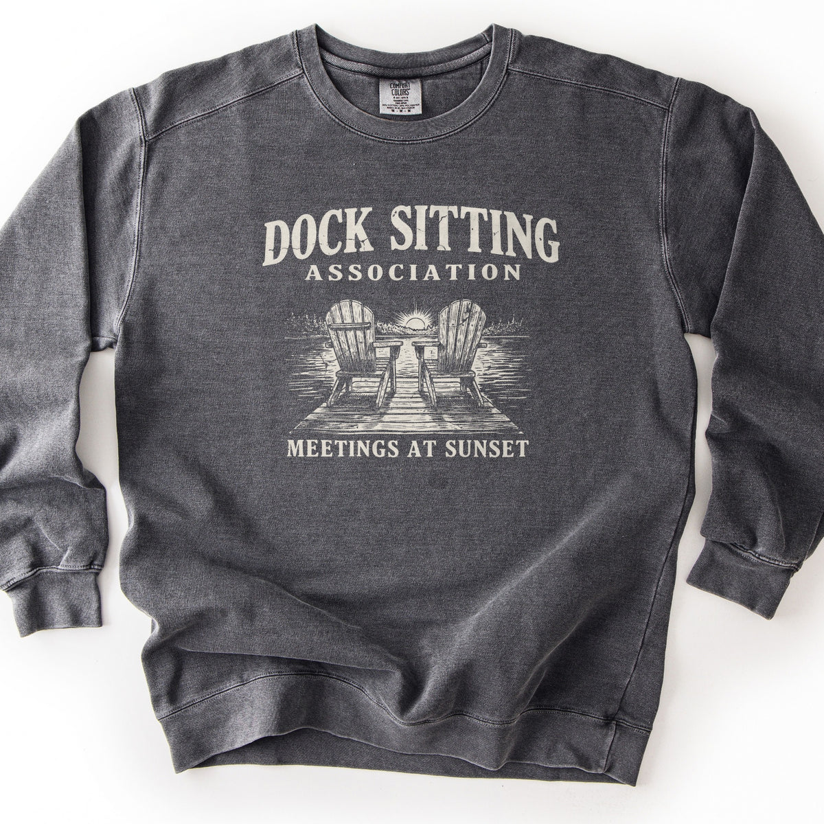 Dock Sitting Association Comfort Colors crewneck sweatshirt in Pepper, laid flat. Cream print: Dock Sitting arched over Association, a sketch of two Adirondack chairs on a wooden dock facing a lake at sunset, and Meetings at Sunset below. Shown on a white background.