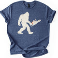 Lumber Squatch Comfort Colors T-shirt in Heather Navy, laid flat. A distressed cream silhouette of Bigfoot walking mid-stride while holding a chainsaw with an orange accent, with no words printed. The shirt has cuffed sleeves and lies on a white background.