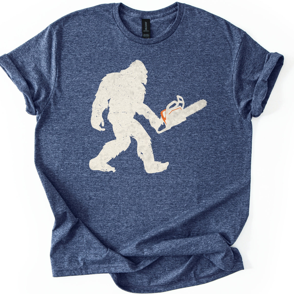 Lumber Squatch Comfort Colors T-shirt in Heather Navy, laid flat. A distressed cream silhouette of Bigfoot walking mid-stride while holding a chainsaw with an orange accent, with no words printed. The shirt has cuffed sleeves and lies on a white background.