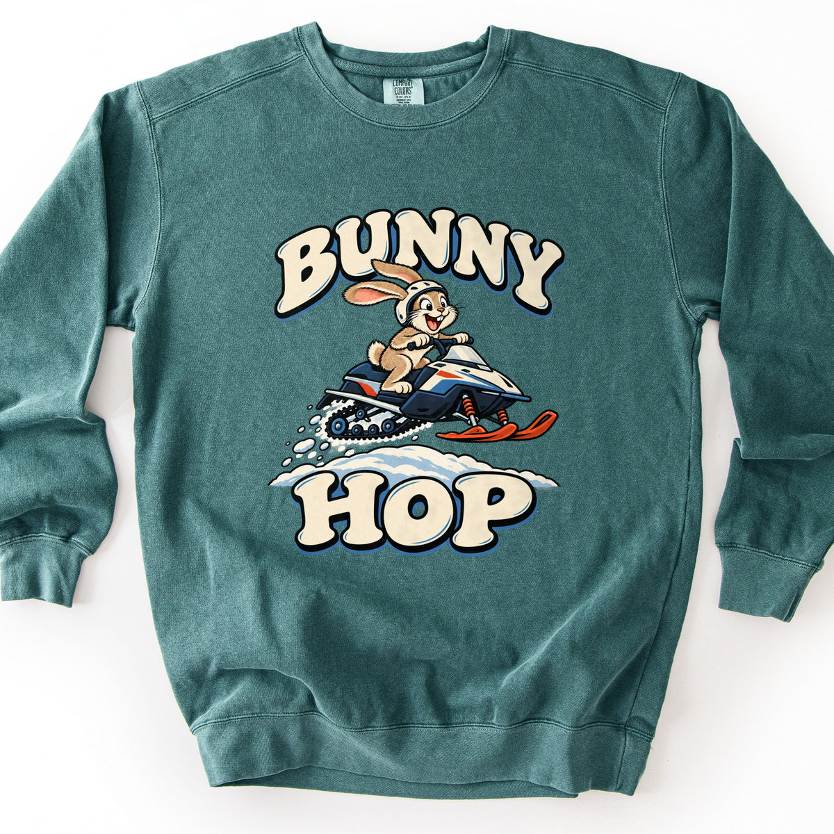 Bunny Hop Comfort Colors crewneck sweatshirt in Blue Spruce, laid flat. A cartoon rabbit in a white helmet rides a navy, white and orange snowmobile over a snowbank, between the words Bunny Hop in cream bubble letters. The sweatshirt lies on a plain white background.
