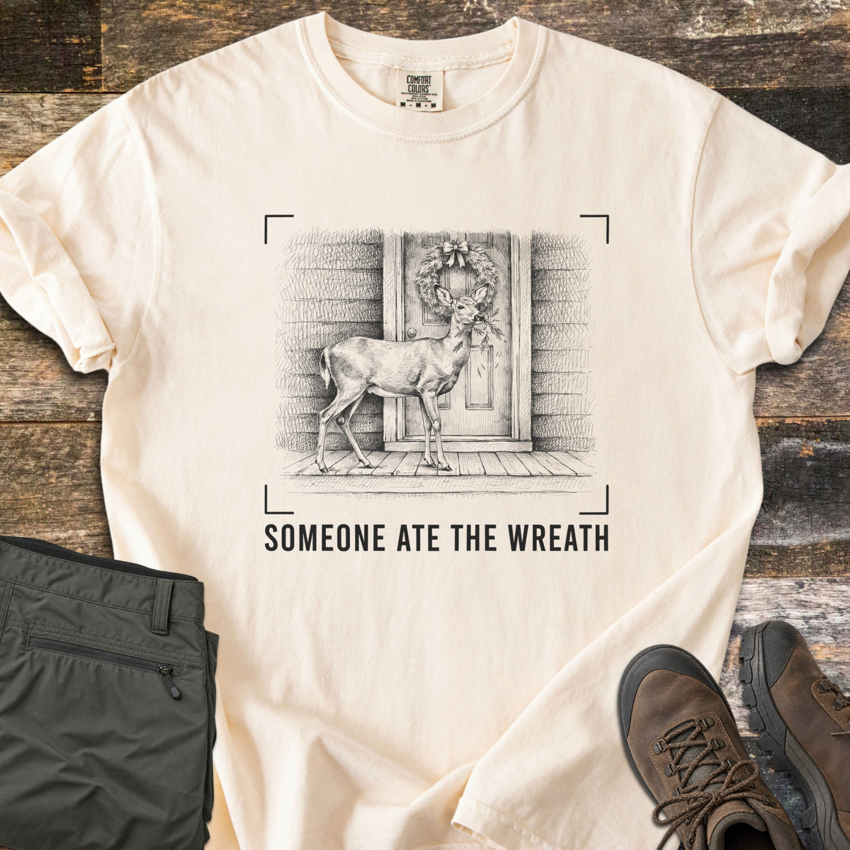 Someone Ate The Wreath Comfort Colors T-shirt in Ivory, laid flat. A sketched deer nibbles a wreath on a cabin's front door, framed by corner marks above the words Someone Ate The Wreath. Shown on rustic wood planks with folded olive pants and brown hiking boots.