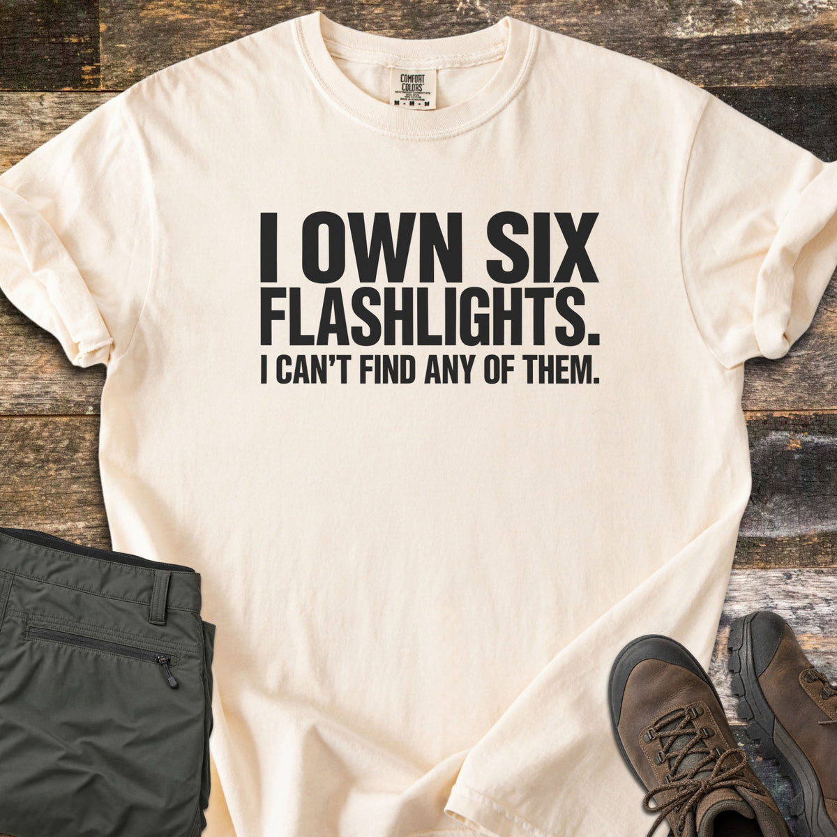 Six Flashlights Comfort Colors T-shirt in Ivory, laid flat. Bold black text reads I OWN SIX FLASHLIGHTS. I CAN'T FIND ANY OF THEM. The shirt has rolled sleeves and lies on weathered wood beside folded olive cargo pants and brown hiking boots.
