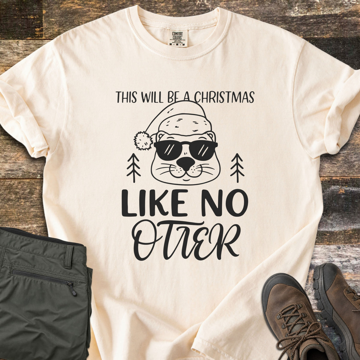 Christmas Like No Otter Comfort Colors T-shirt in Ivory, laid flat. A cartoon otter in a Santa hat and sunglasses sits between two pine trees, with This Will Be a Christmas above and Like No Otter below, in black. Olive cargo pants and brown hiking boots lie on weathered wood.