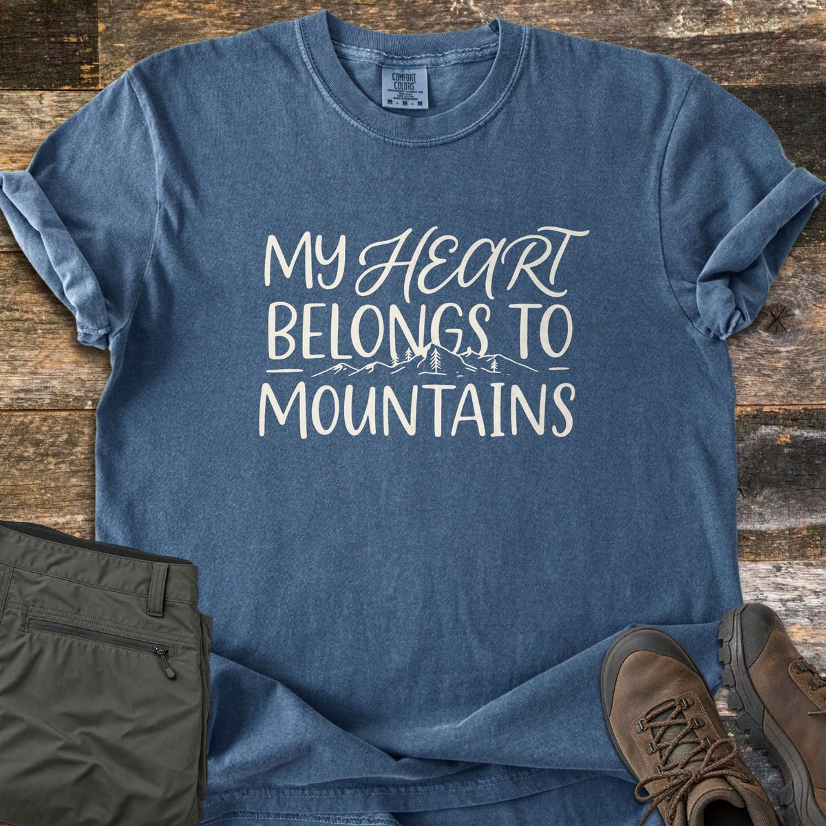 My Heart Belongs to Mountains Comfort Colors T-shirt in Denim, laid flat. Cream lettering reads My Heart Belongs to Mountains, with a line of mountain peaks and pine trees beneath. It lies on rustic wood planks beside olive hiking pants and brown hiking boots.