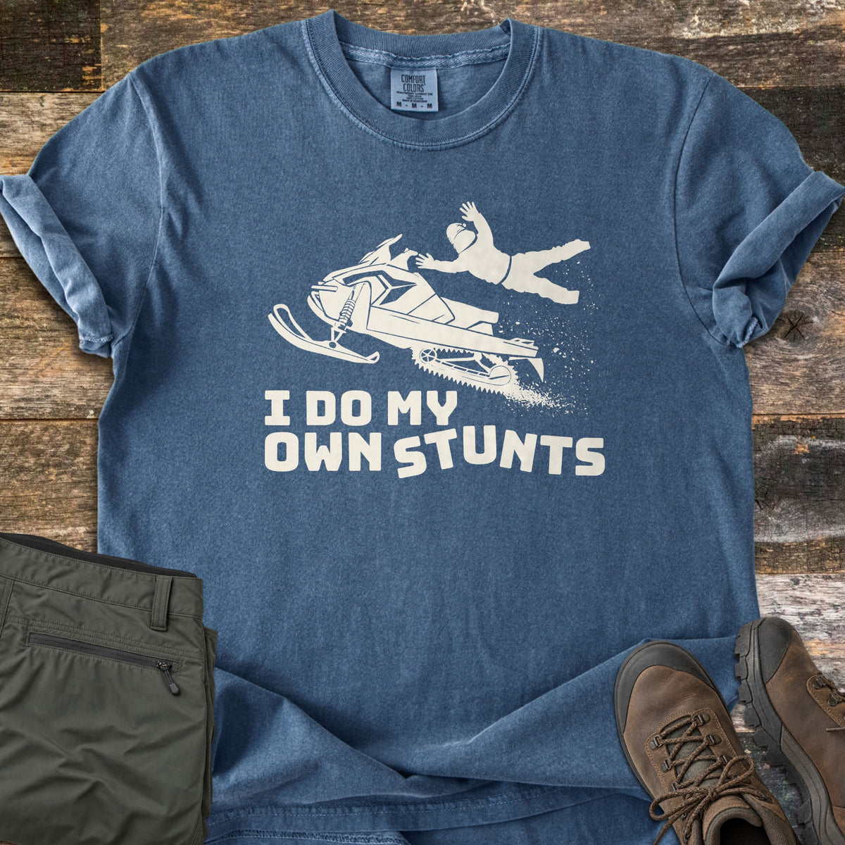 I Do My Own Stunts Comfort Colors T-shirt in Denim, laid flat. A cream silhouette of a rider flying off a jumping snowmobile, snow spraying behind, above the words I Do My Own Stunts. Folded olive cargo pants and brown hiking boots sit beside it on weathered wood.