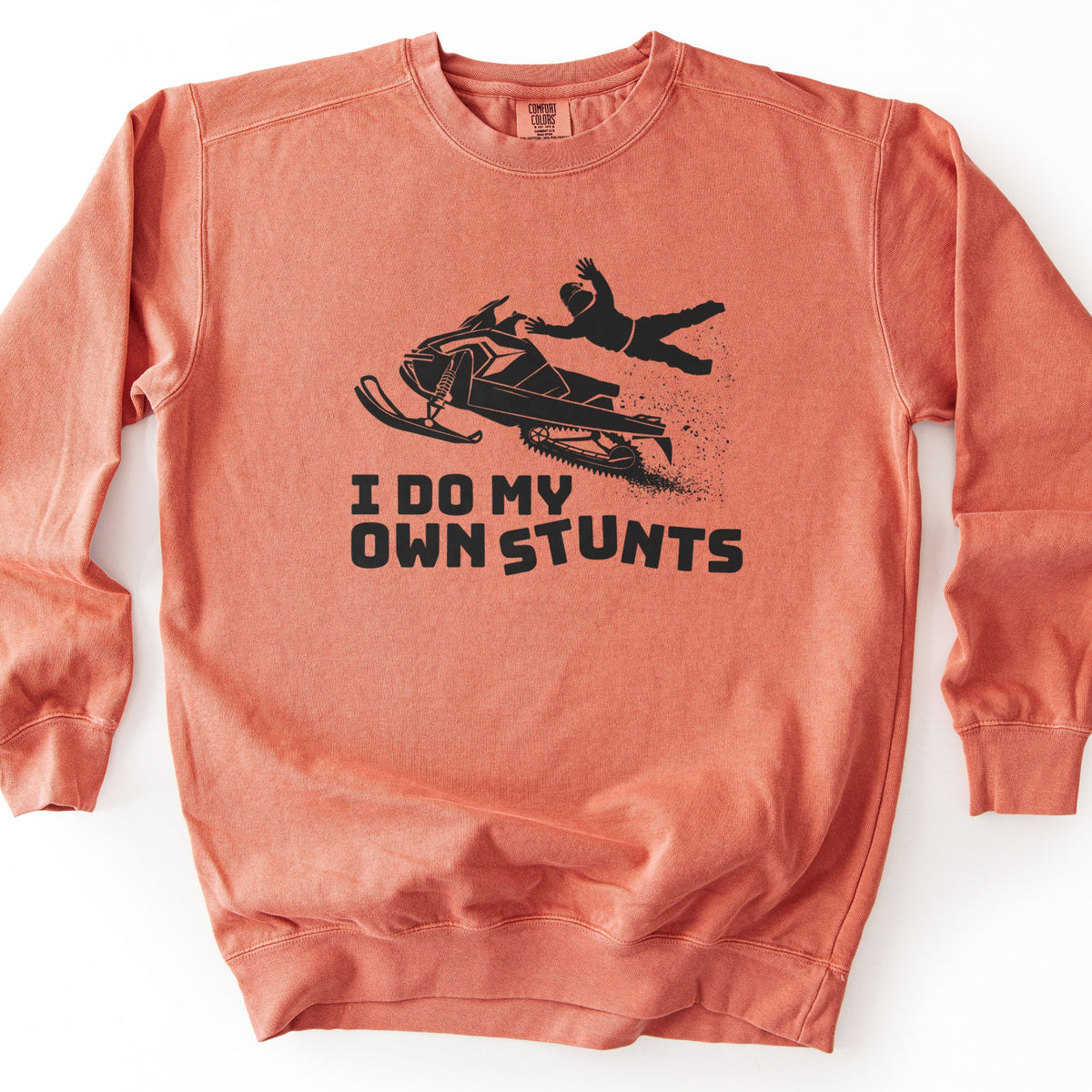 I Do My Own Stunts Comfort Colors crewneck sweatshirt in Terracotta, laid flat. A black print shows a helmeted rider thrown from an airborne snowmobile with snow spraying behind, above the words I DO MY OWN STUNTS. The sweatshirt lies on a white surface.