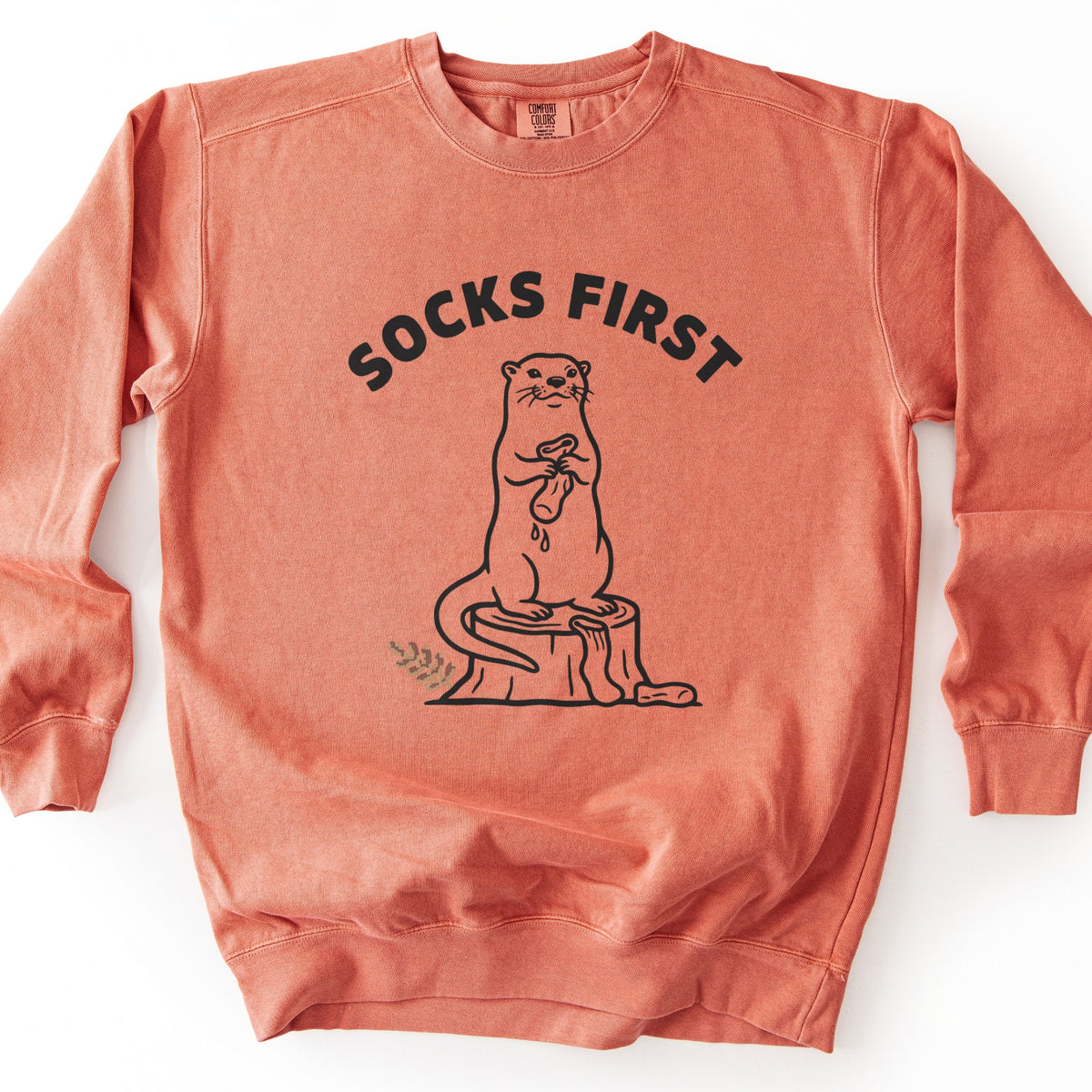 Socks First Comfort Colors crewneck sweatshirt in Terracotta, laid flat. A black line drawing of an otter on a tree stump wringing a dripping sock, with another sock at the stump's base, under the arched words Socks First. A small fern sprig is at left. White background.