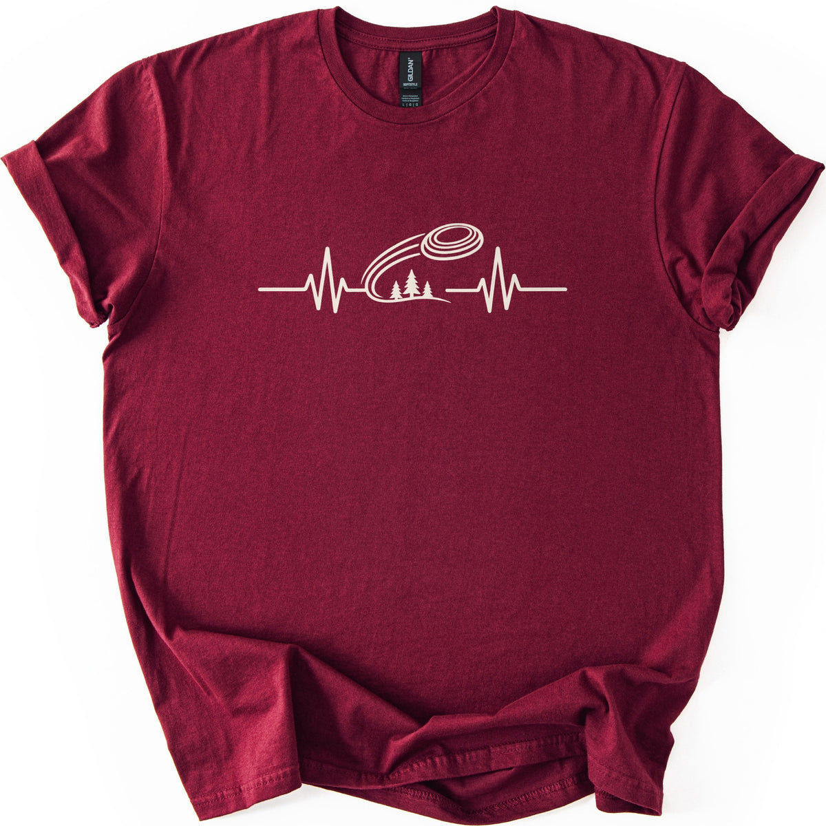 Trap Shooting Heartbeat Comfort Colors T-shirt in Maroon, laid flat. A cream heartbeat line with a clay target flying above three pine trees on a hill, with no words. The shirt lies on a white background with its sleeves rolled and the hem loosely folded.